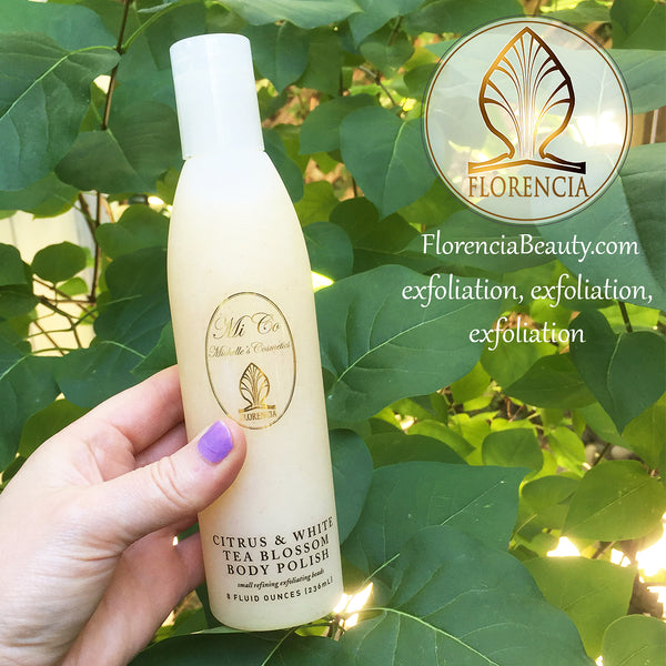 A hand holding a bottle of Citrus & White Tea Blossom Body Polish with greenery in the background. 